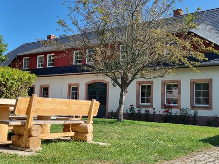 Außenansicht des Jugendbegegnungszentrums Seelitz mit Innenhof, heller Gebäudefassade und einer Holzsitzgarnitur in der Mitte, umgeben von Rasen und Pflanzen bei sonnigem Wetter.