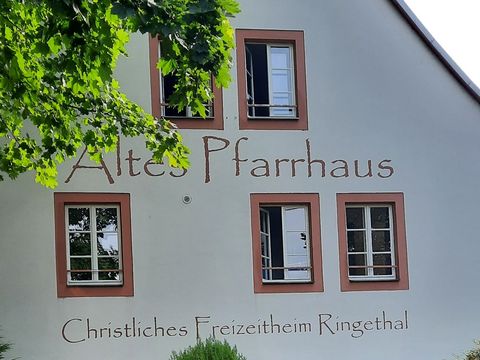 Außenansicht des Alten Pfarrhauses in Ringethal, ein zweigeschossiges Haus mit heller Fassade und Satteldach, umgeben von Wiese und Bäumen bei Tageslicht.