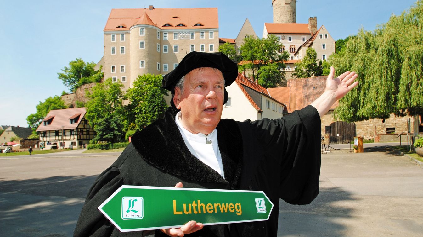 Person verkleidet als Martin Luther, hält einen Wegweiser „Lutherweg Sachsen“ in der Hand, stehend vor Burg Gnandstein.