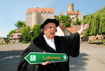 Person verkleidet als Martin Luther, hält einen Wegweiser „Lutherweg Sachsen“ in der Hand, stehend vor Burg Gnandstein.