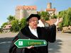 Person verkleidet als Martin Luther, hält einen Wegweiser „Lutherweg Sachsen“ in der Hand, stehend vor Burg Gnandstein.