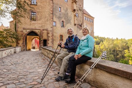 Das Bild zeigt ein Pärchen Wanderer, die vor der Burg Kriebstein auf der Mauer der Burgbrücke eine Rast einlegen..