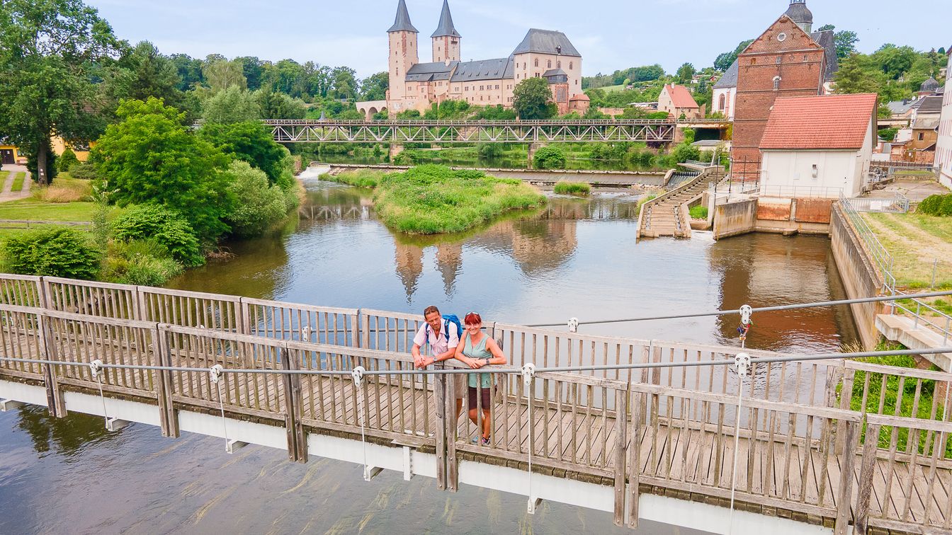 Die Drohnenaufnahme zeigt die Hängebrücke mit zwei Wanderern vor der imposnaten Kulisse von Mulde, Schloss und Kirche.
