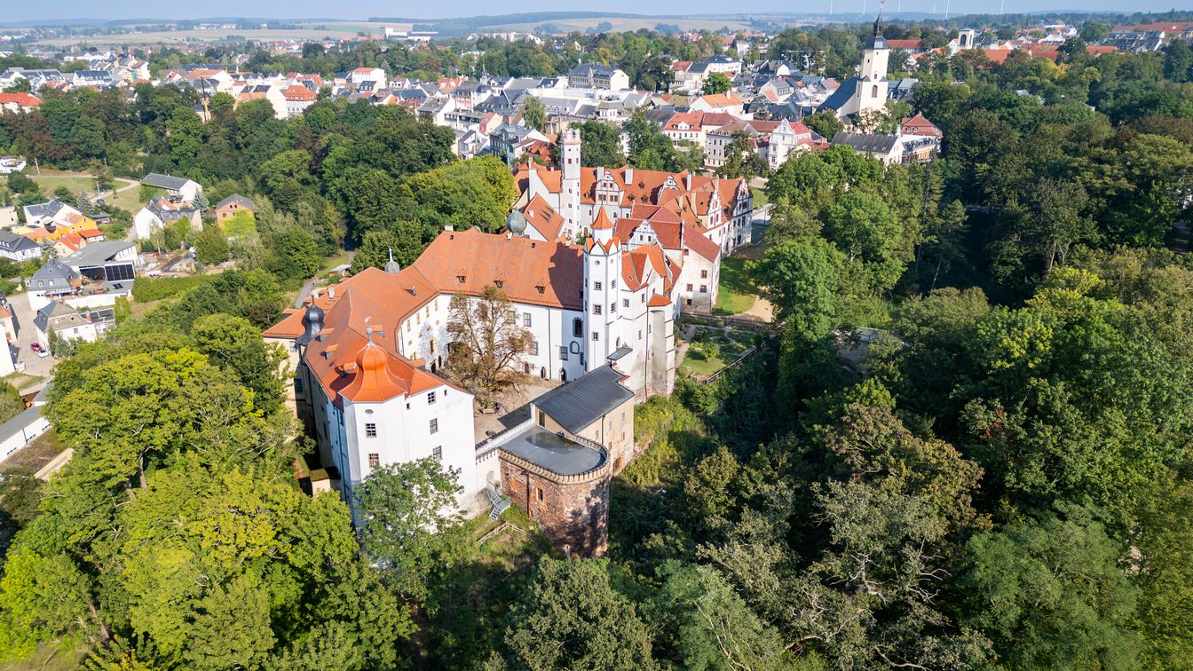Schloss Glauchau, ein großes Renaissancebauwerk, mit zwei verbundenen Schlossflügeln, roten Dächern und hellen Fassaden. Das Schloss steht leicht erhöht über der Stadt Glauchau und ist von Grünflächen und Bäumen umgeben.