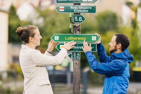 2 Wegewarte befestigen das Schild des Lutherwegs Sachsen an einem Pfosten.