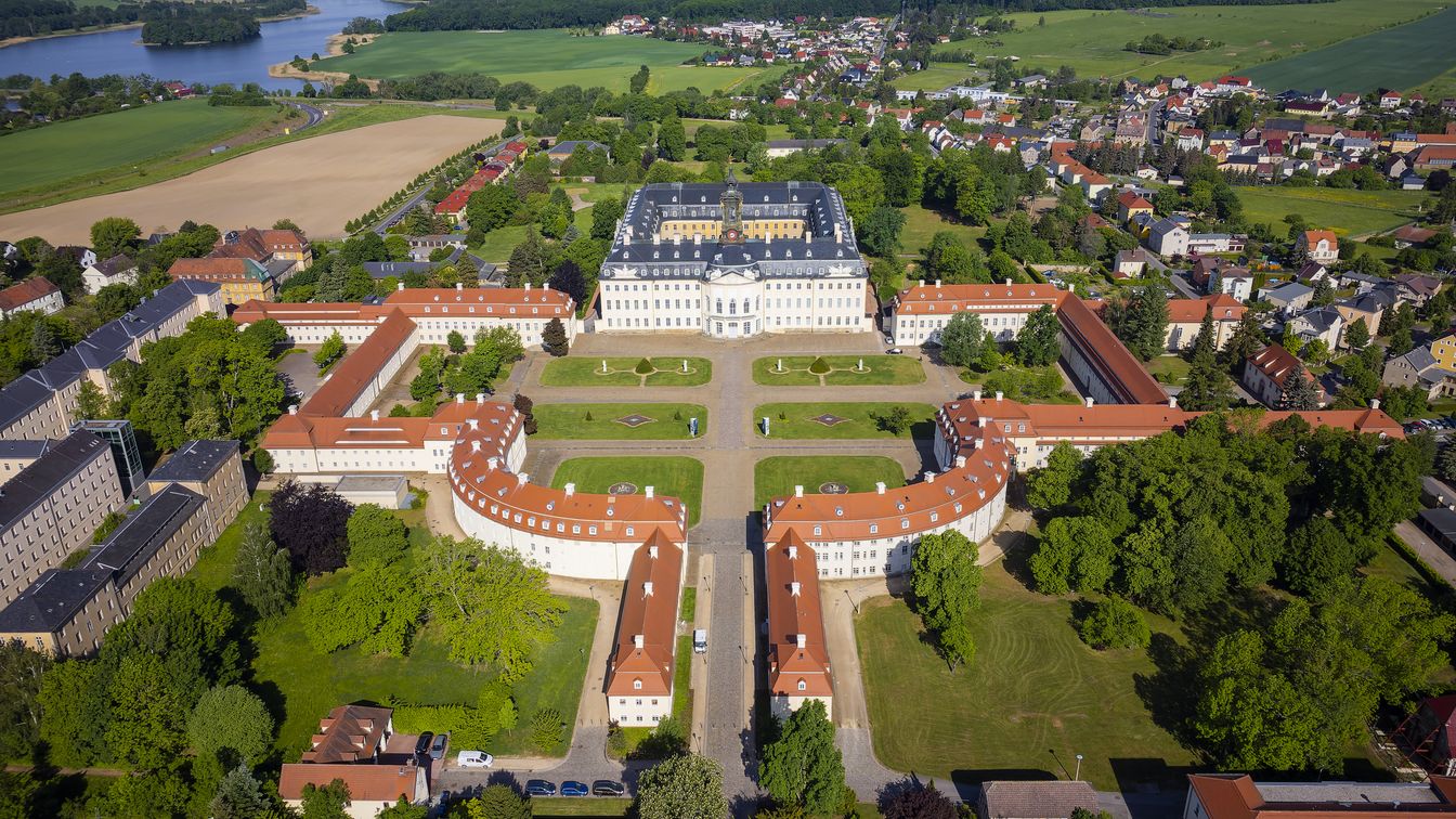 Das Bild zeigt das Schloss Hubertusburg in Wermsdorf als Drohnenaufnahme, umgeben von Wald- und Teichlandschaften. 