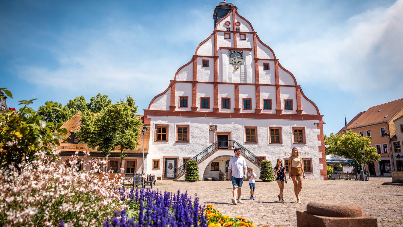 Das Bild zeigt eine junge Familie auf dem Marktplatz vor dem markant rot weißen Rathaus.